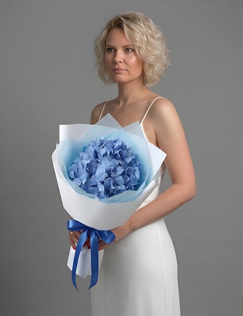 A single, large, and beautiful blue hydrangea bloom from Bloom & Art set against a neutral background, highlighting its dense, cloud-like petal structure.